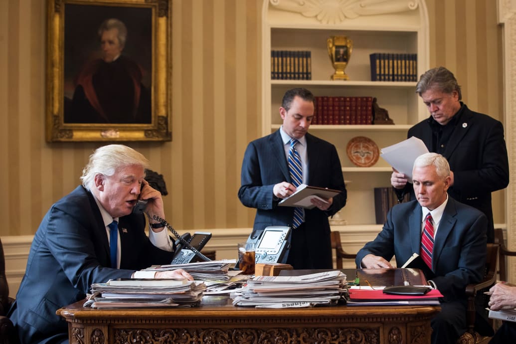 Donald Trump speaks on the phone in the Oval Office.  Mike Pence is sitting in a chair facing him and in the background Steve Bannon, dressed all in black, is holding a piece of paper.