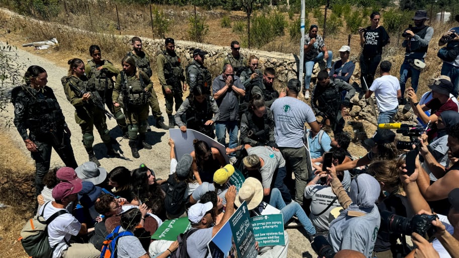 A line of Israeli soldiers faces a group of pro-Palestinian protesters, some sitting and some standing in a dirt road holding signs.