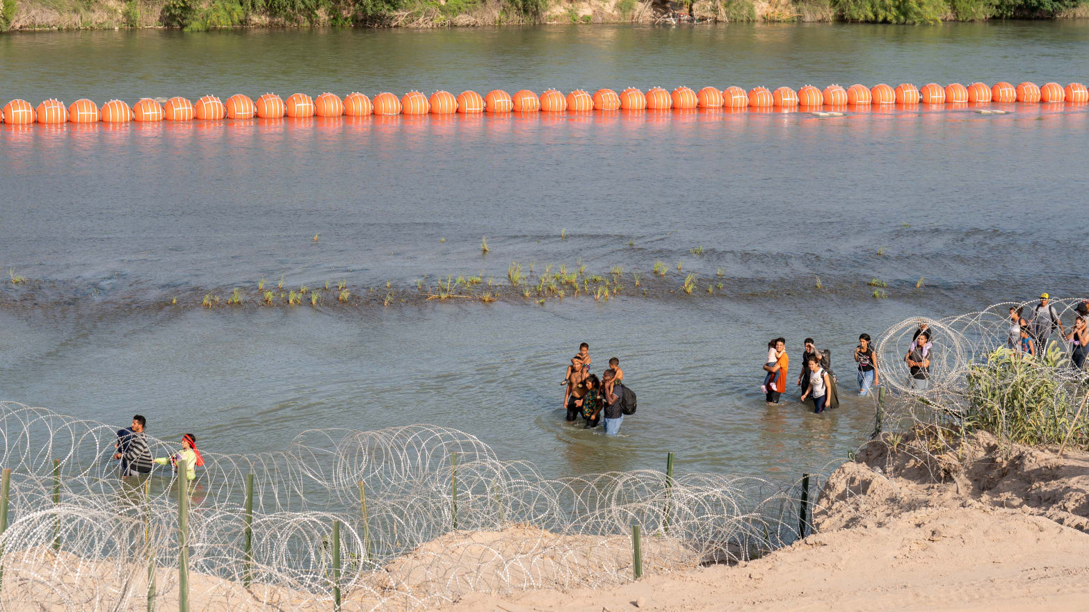 Migrants walk by a string of buoys placed on the water along the Rio Grande border with Mexico in Eagle Pass, Texas.
