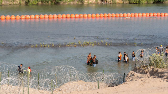 Migrants walk by a string of buoys placed on the water along the Rio Grande border with Mexico in Eagle Pass, Texas.