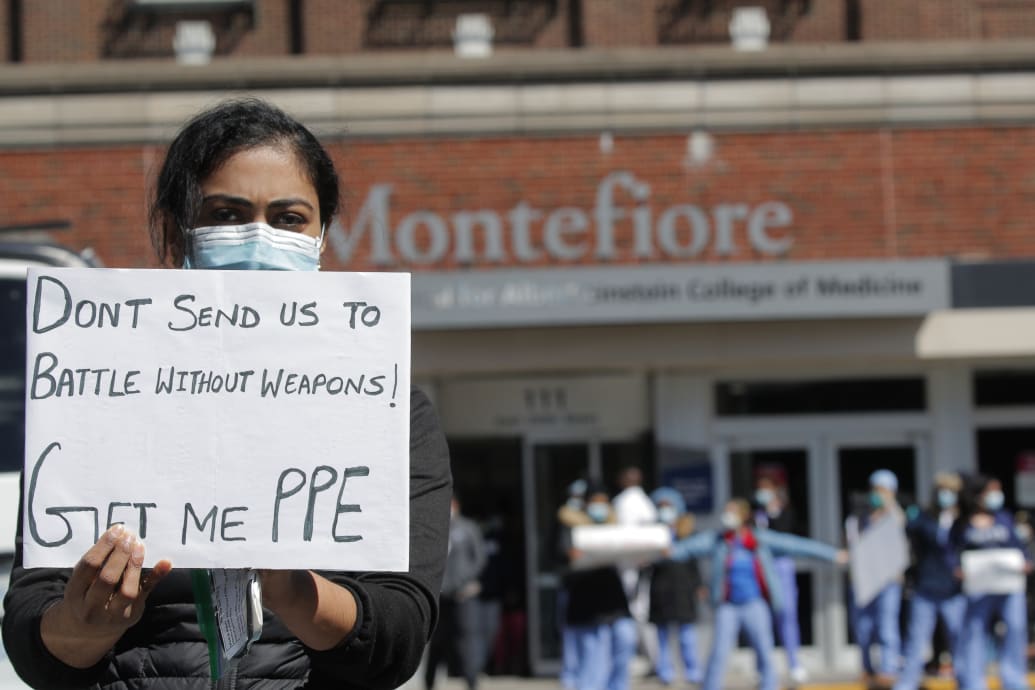 A woman in a mask in front of a hospital holding a sign: Don’t send us to battle without weapons! Get me PPE.