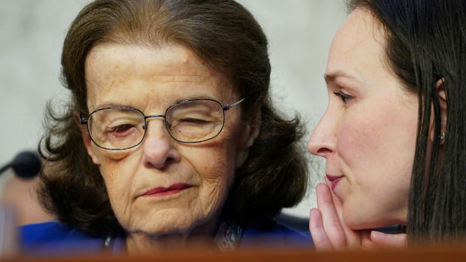 A picture of U.S. Senator Dianne Feinstein (D-CA) listening to an aide. She was recently reminded to “just say aye” when asked to vote on the defense appropriations bill.