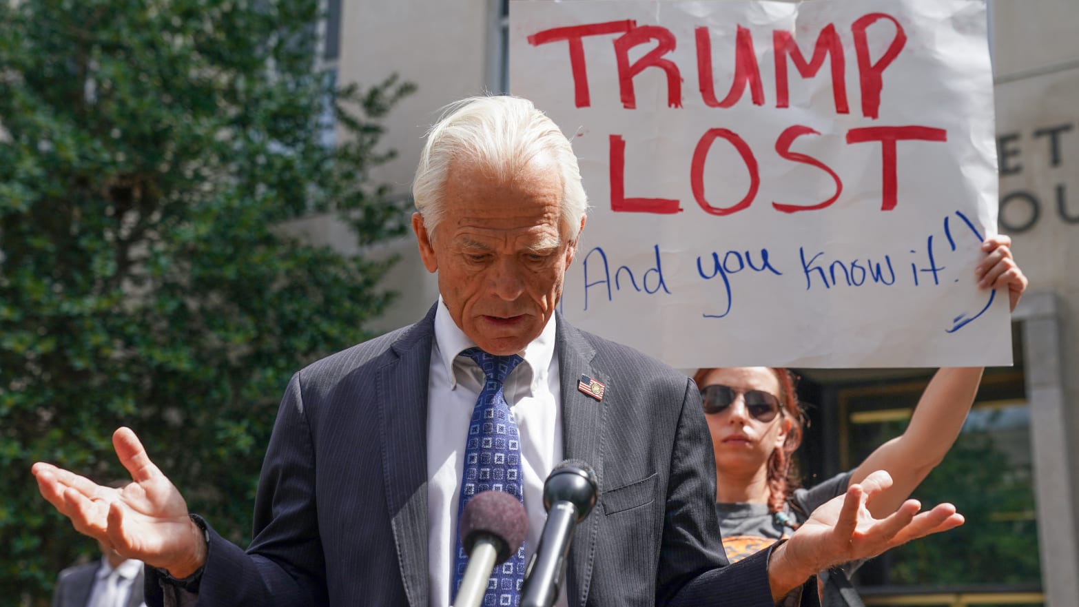 Peter Navarro, wearing a suit, stares down during a press conference where he was heckled by a protester holding a “TRUMP LOST” sign.