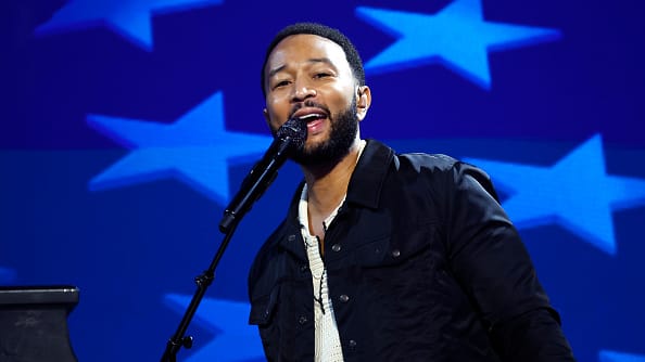 John Legend stands at a microphone, a bright blue backdrop of lighter blue stars behind him