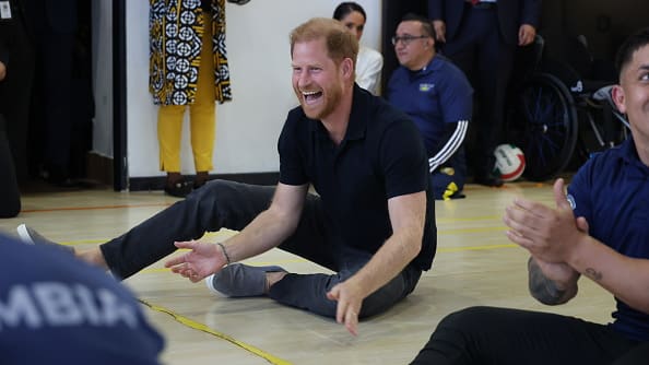 Prince Harry laughs while sitting on the floor of what looks like a gymnasium, people clapping around him.