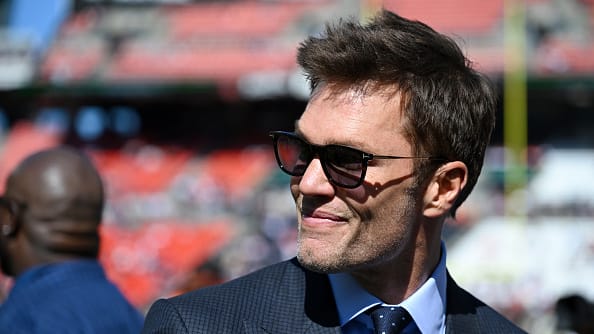 Tom Brady, wearing a suit and sunglasses, smiles as he looks to the left on Huntington Bank Field in Cleveland, Ohio. 