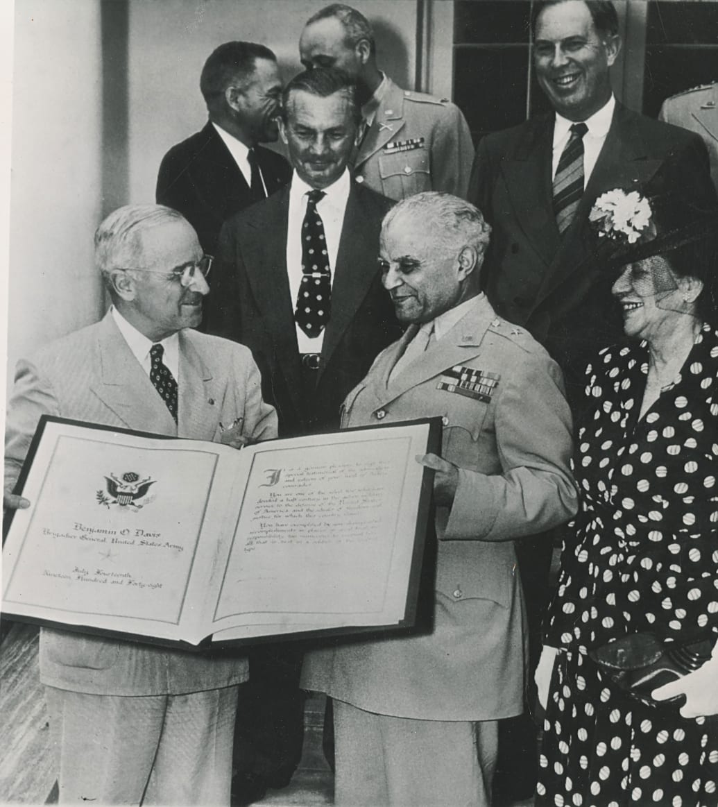 An archival photo of President Truman presenting Ollie with a scroll surrounded by other politicians. 