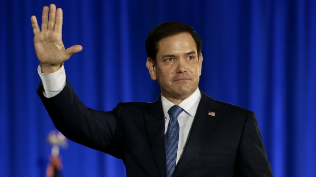 Sen. Marco Rubio walks across a stage with a blue backdrop, raising one hand