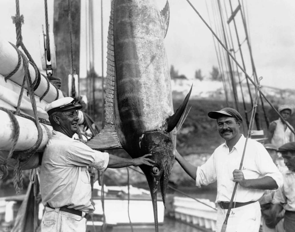 Ernest Hemingway stands with a fishing rod and a marlin, while Captain Joe Russell from Key West looks on.