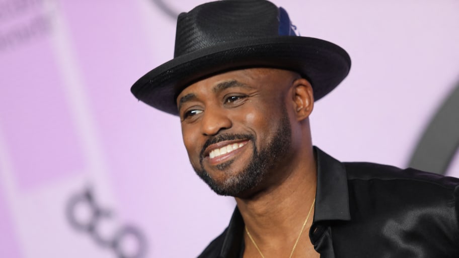 Wayne Brady, wearing a hat and black shirt, poses and smiles at an awards show.