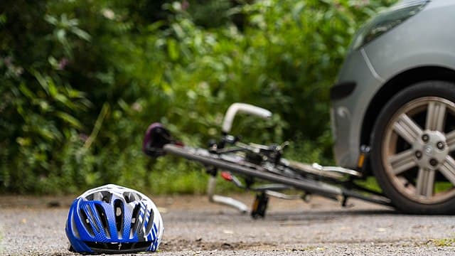 Photo of a helmet lying on ground with bicycle trapped underneath a car in the background during a road traffic accident involving a bike crash.