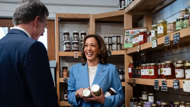 Kamala Harris holding two jars of food and talking to a man with his back to camera in an artisanal food store