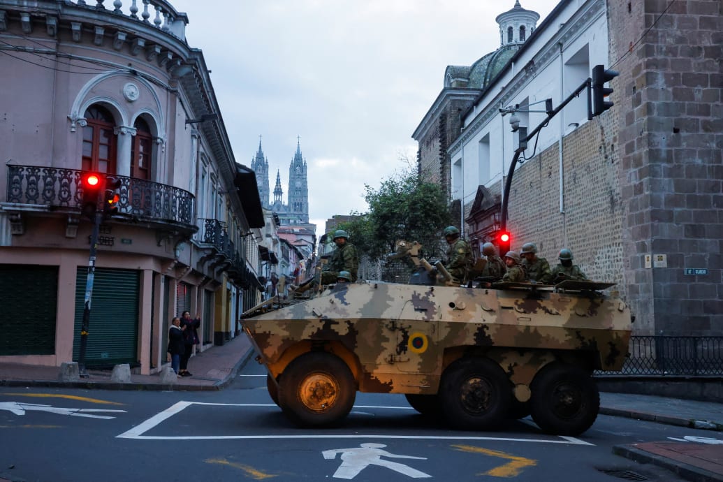 A military vehicle with soldiers inside drives down a street in the historic center of Quito, with a cathedral in the background.