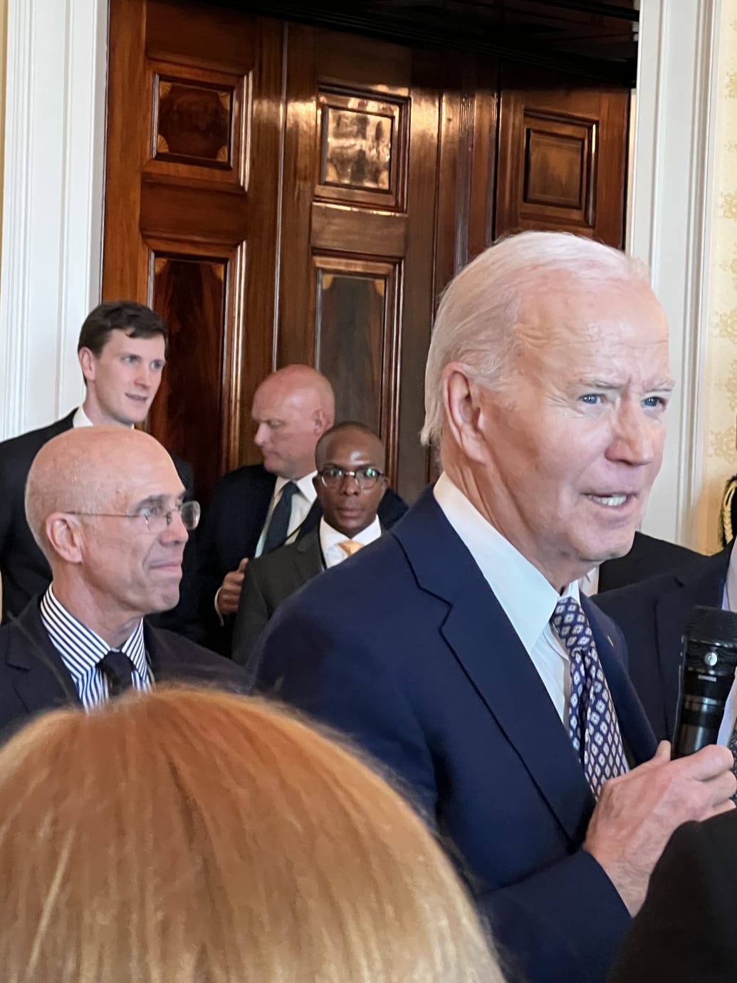 Joe Biden speaking into a microphone with Jeffrey Katzenberg behind him, in a White House room.