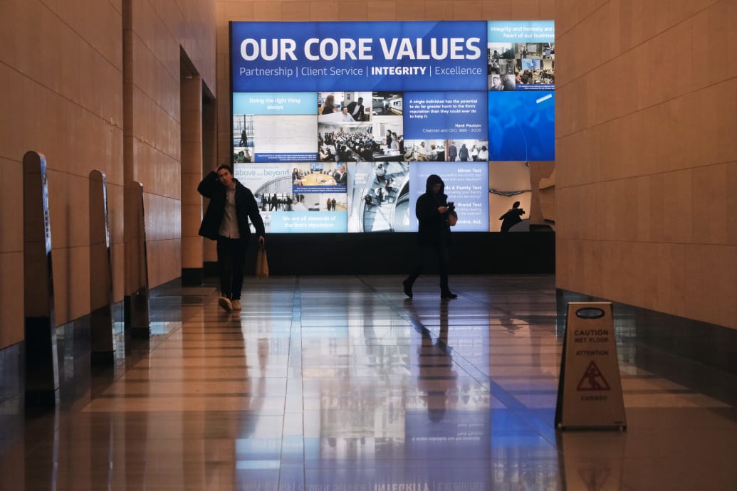 People walk through the lobby of the Goldman Sachs headquarters; an illuminated ad on the wall reads: “Our Core Values.”