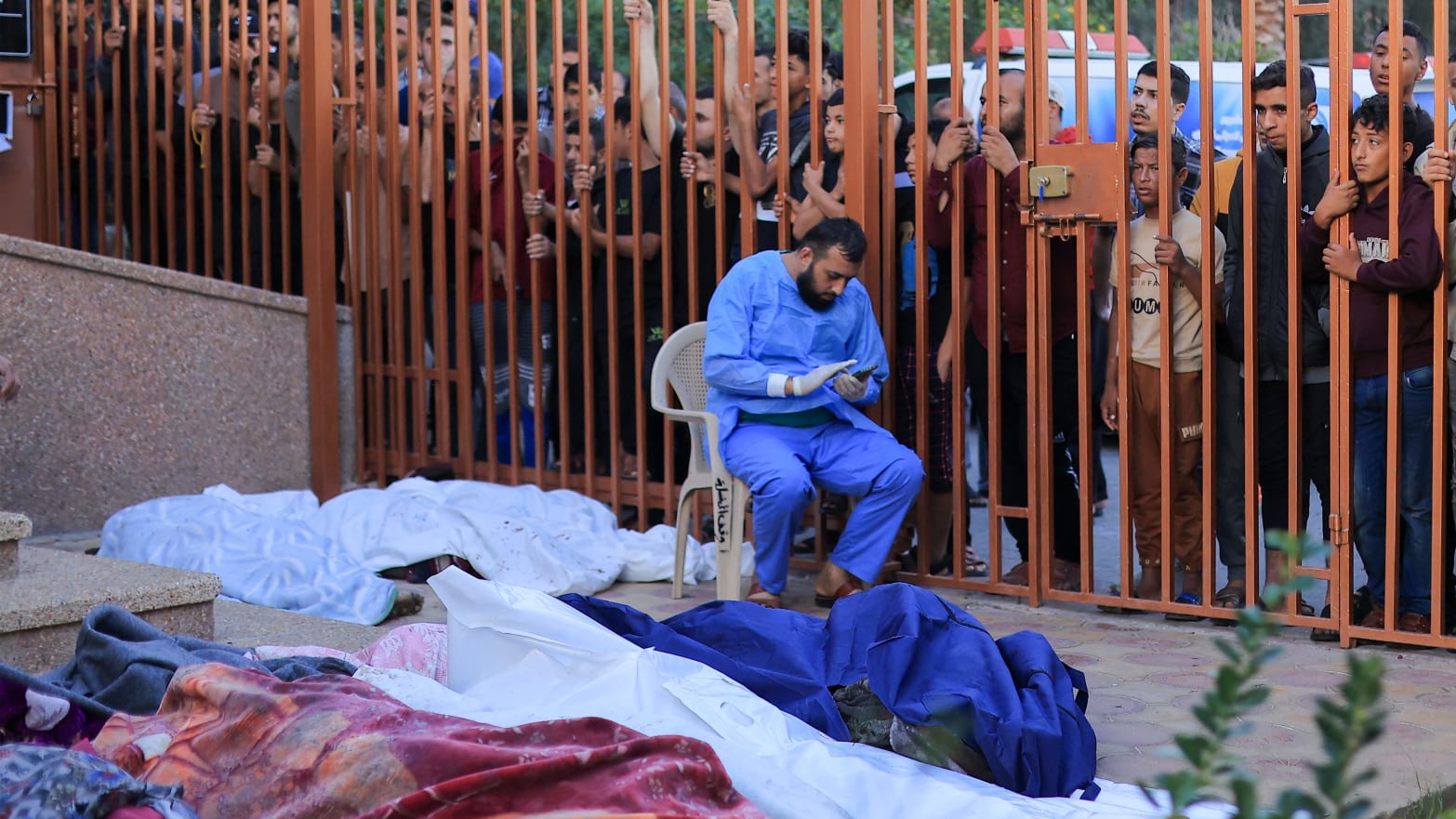 A medical worker sitting among bodies covered in white shrouds, with people looking over through a gate.