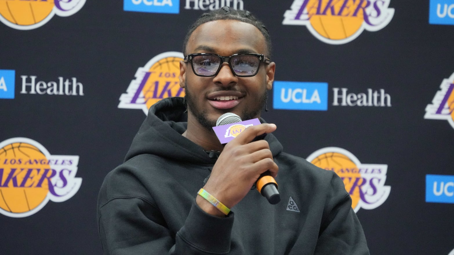 Bronny James smiles while holding a microphone at a Lakers press event.