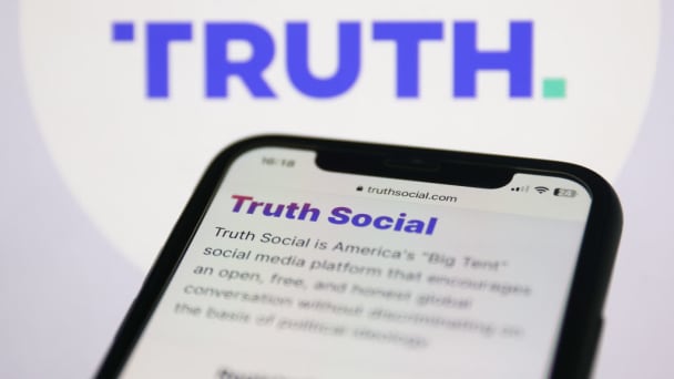 Truth Social website is seen displayed on a phone screen and the Truth Social logo is displayed on a screen behind it. 