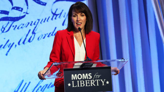 Tina Descovich, wearing red, speaks on stage behind a podium that has a “Moms For Liberty” sign.