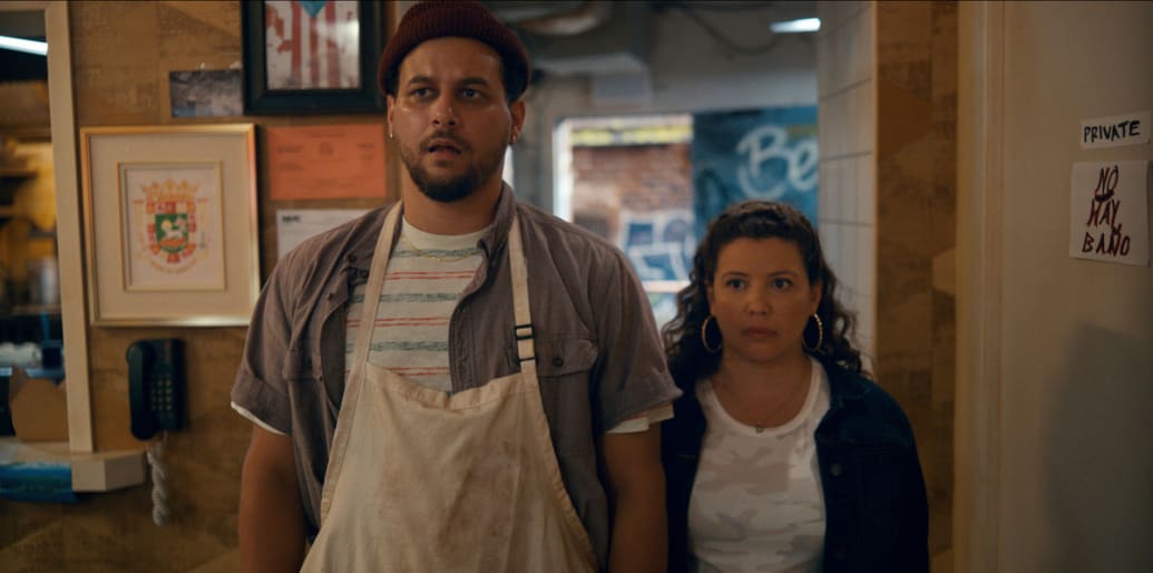Justina Machado and Alejandro Hernandez stand next to each other in a a still from 'The Horrors of Dolores Roach'