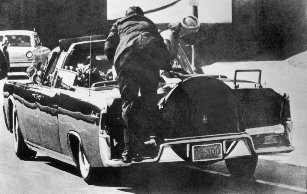 Just after John F. Kennedy was shot, Jacqueline Kennedy stands up in the presidential car to lift up the body of her husband. In the foreground, a Secret Service agent jumps onto the back fender of the car.