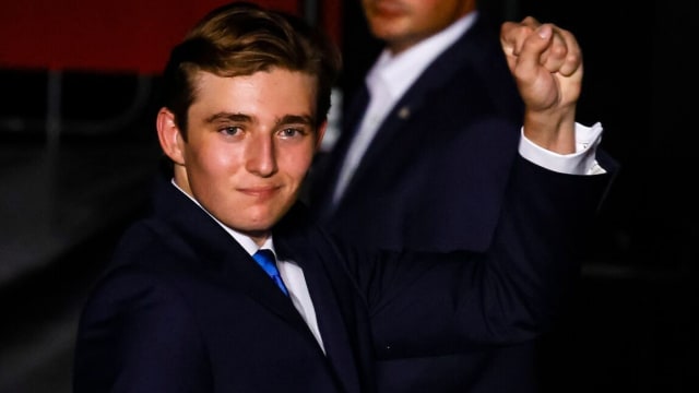 Barron Trump fist pumps while standing and wearing a blue suit, white shirt and blue tie