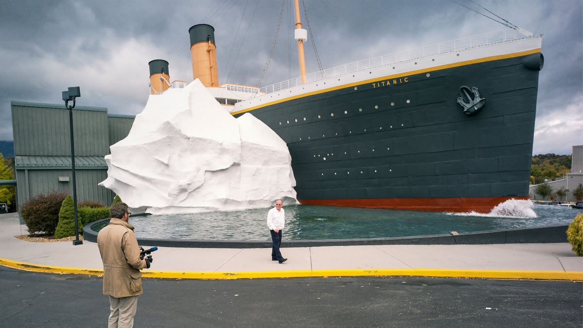 A still from “How to with John Wilson” that shows John WIlson in the foreground looking at a replica of the Titanic