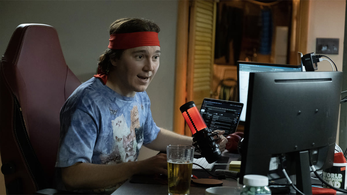 A still from “Dumb Money” shows Paul Dano at a computer setup with a microphone in front of him.