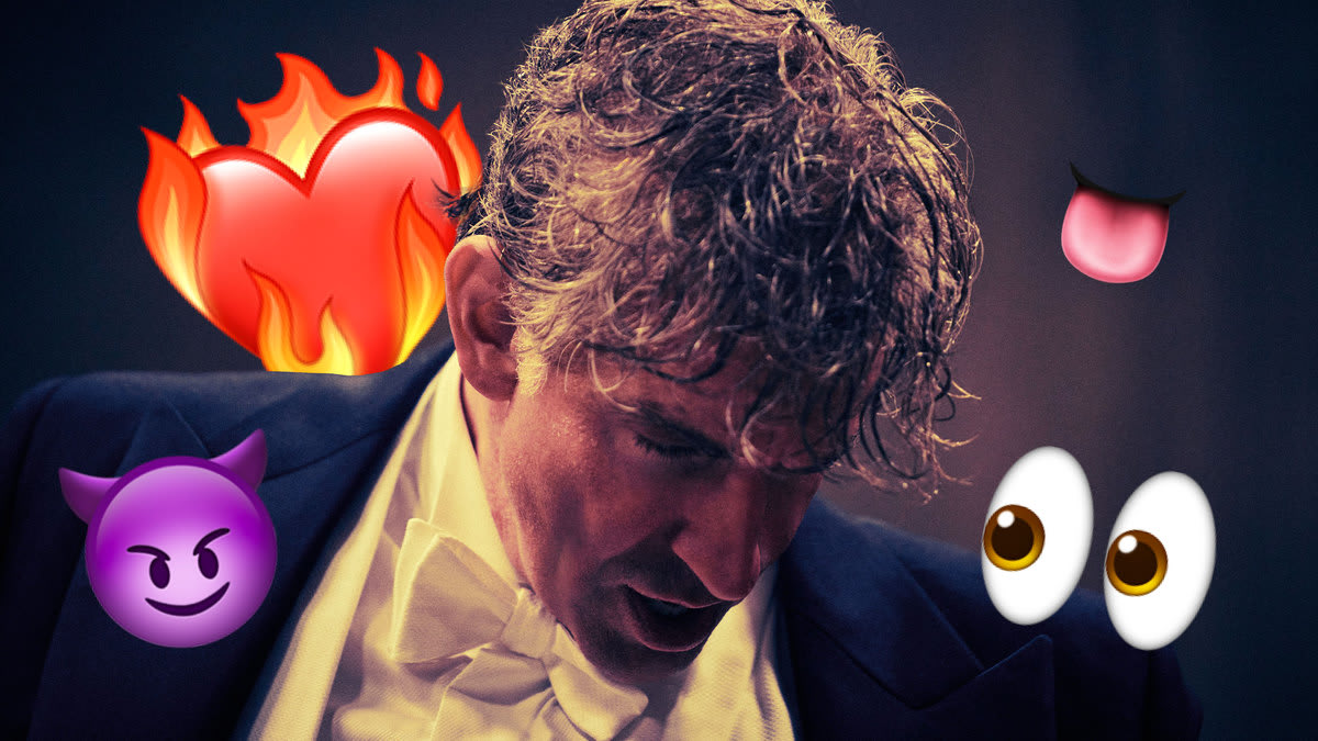 A close up photo of Bradley Cooper as Leonard Bernstein in a conductor’s tuxedo looking down while flirty emojis surround him.