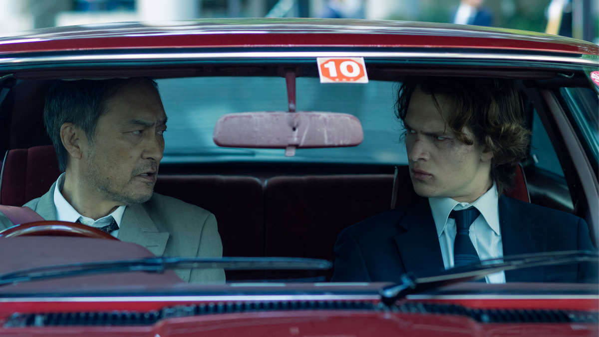 Ken Watanabe and Ansel Elgort inside of a car looking at each other in a still from ‘Tokyo Vice’
