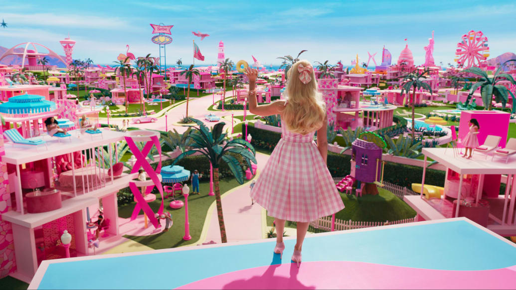 Margot Robbie as Barbie stands waving on the roof of her home, looking out at the bright pink world before her.
