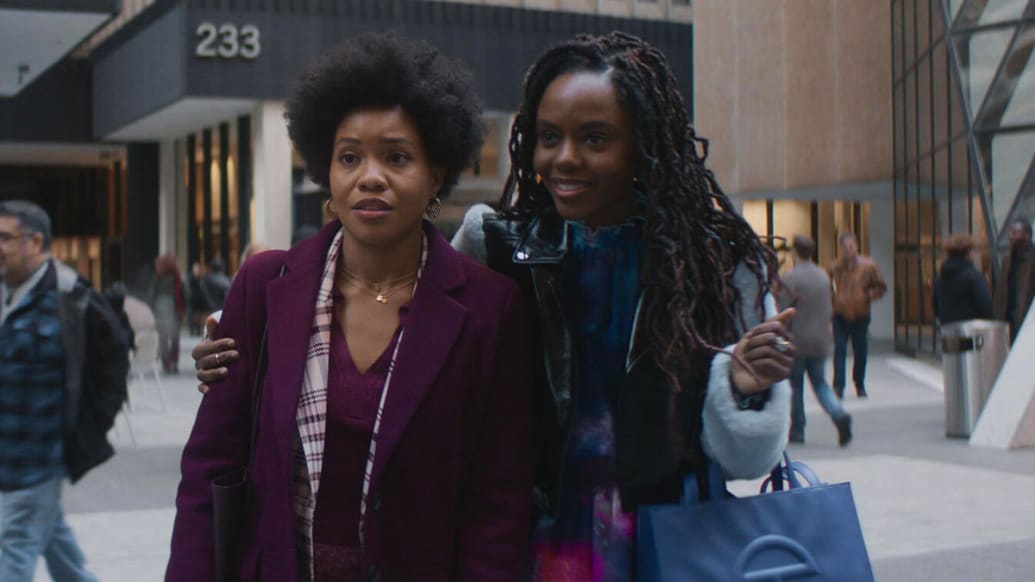 Two young Black women stand outside their office building, close to each other.