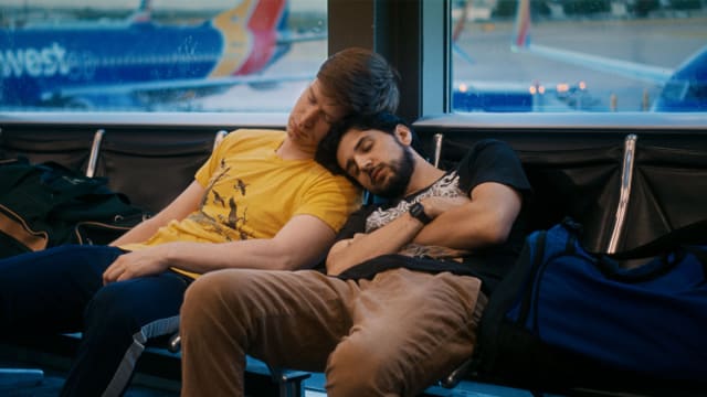 Jacob Roberts and Ben Treviño lean on each other on seats in an airport in a still from ‘Rent Free’