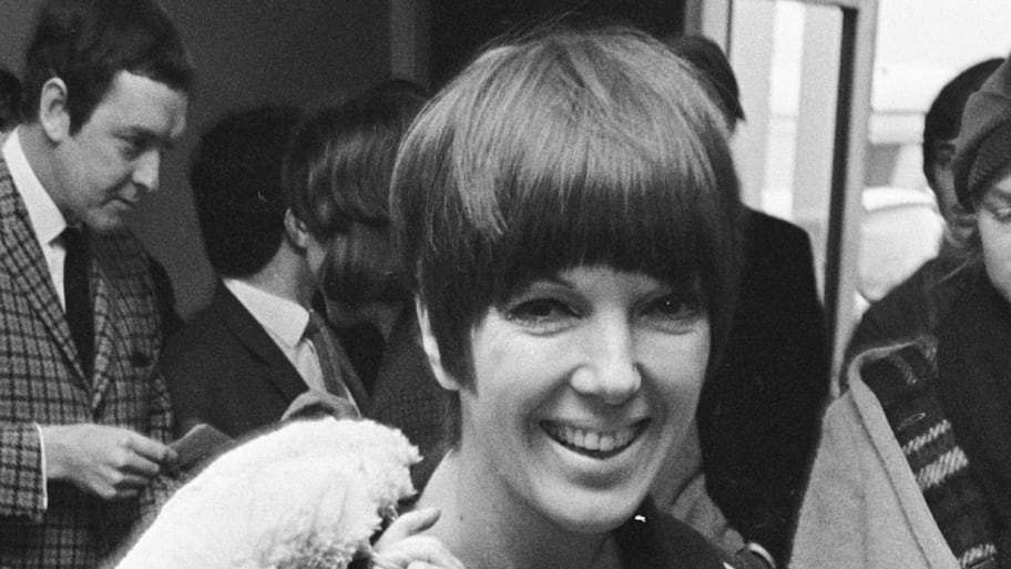Mary Quant wearing a minidress of her own design, with a sheepskin coat and bag thrown over her shoulder, and wearing go-go boots.