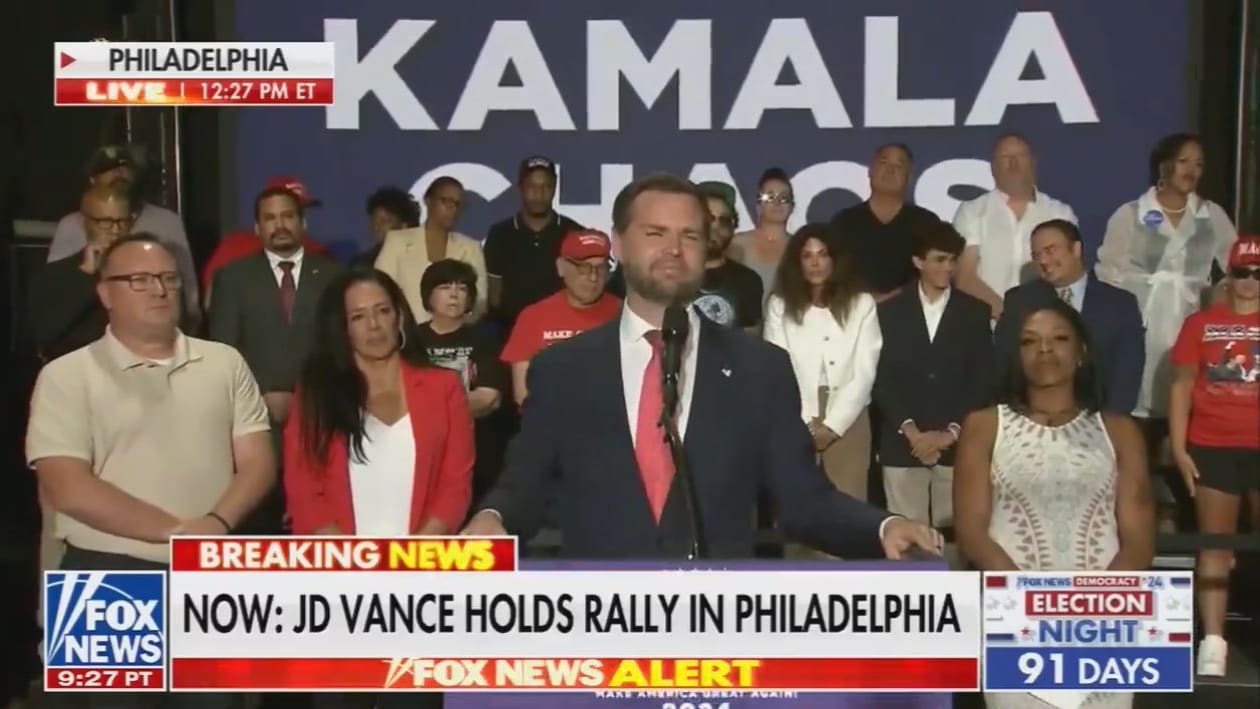 J.D. Vance delivers a speech in front of a crowd and a poster that reads “KAMALA.”