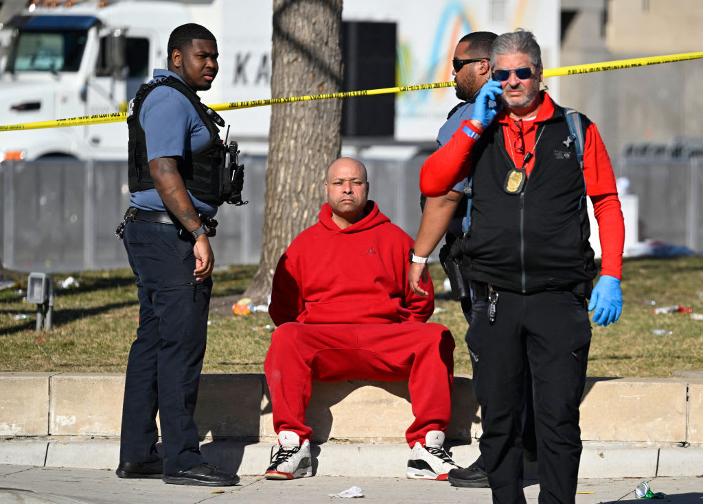 A man, dressed in all red, sits in handcuffed, flanked by cops.