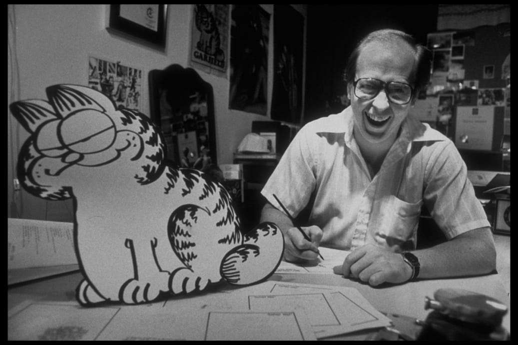 Jim Davis smiles at a desk next to an early drawing of Garfield the Cat.