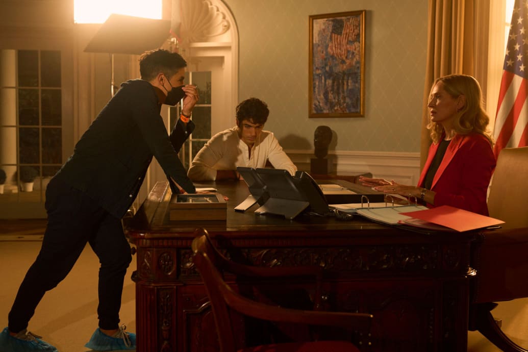 A behind-the-scenes picture of Red, White and Royal Blue that shows Matthew Lopez at a desk talking to Uma Thurman and and Taylor Zakhar Perez.