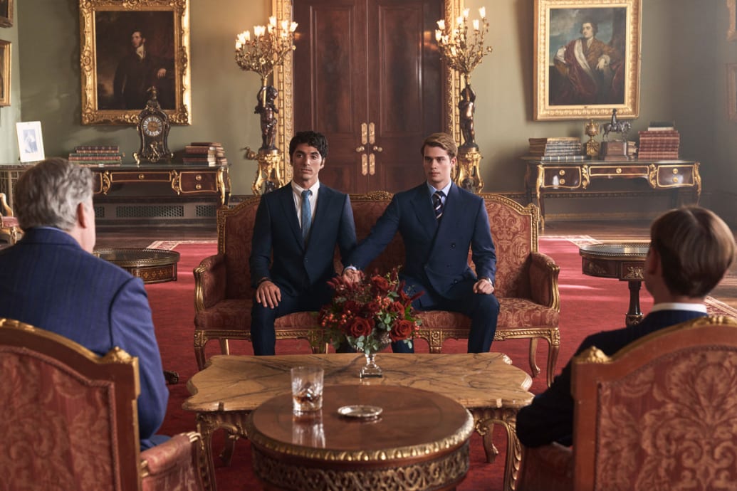 A still of ‘Red, White & Royal Blue’ that shows Nicholas Galitzine as Prince Henry and Taylor Zakhar Perez as Alex Claremont-Diaz holding hands sitting on a couch.
